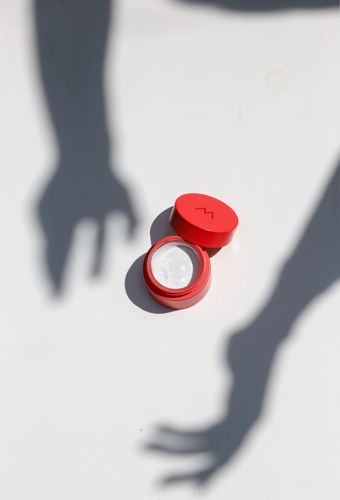 Red-capped jar of Porcelain Hand Balm on a white surface with soft hand-shaped shadows.