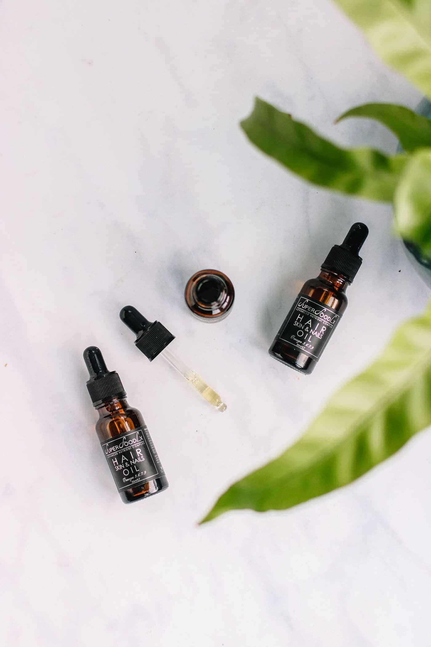 Two amber dropper bottles of Root Revival Oil laid on marble beside fresh green leaves.